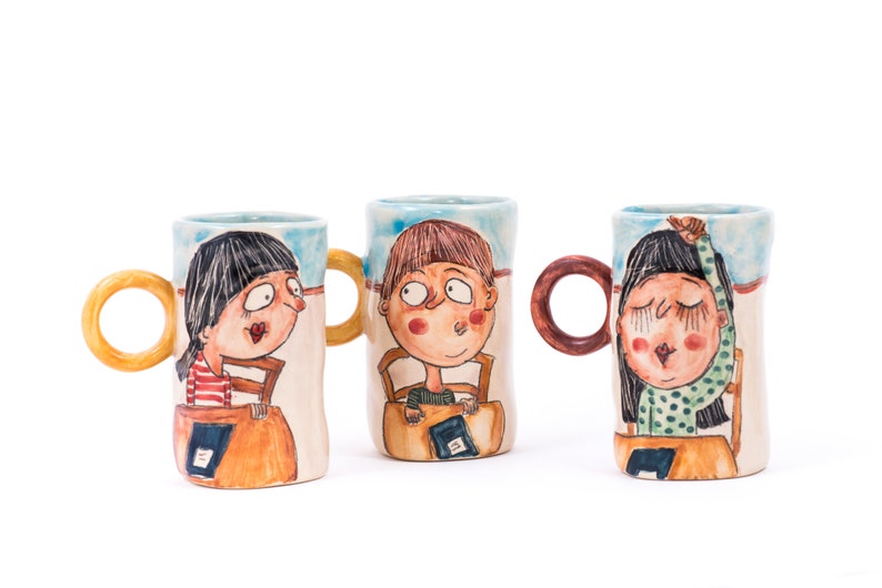 May include: Three ceramic mugs with hand-painted illustrations of children. Each mug features a different child with unique facial expressions and clothing. The mugs have colorful handles and are decorated with a blue and white background.