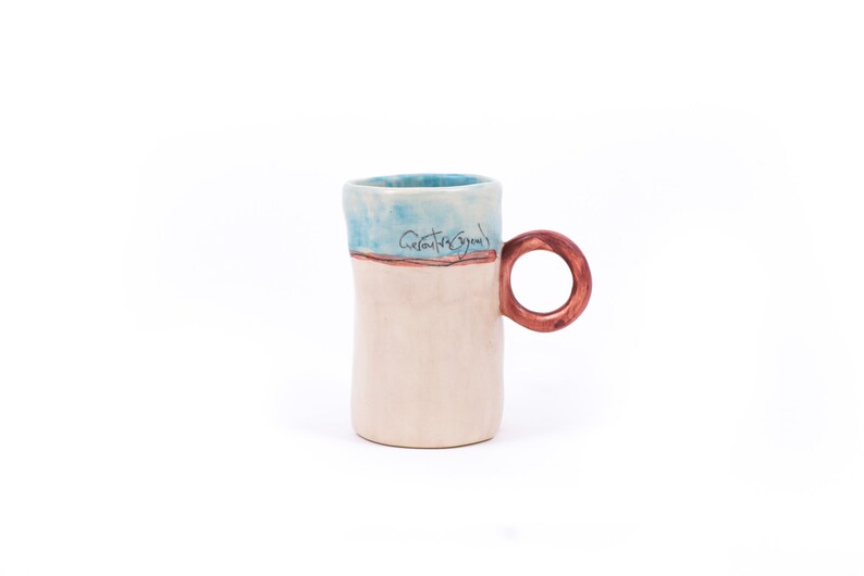 May include: A white ceramic mug with a blue and brown glaze. The mug has a rounded handle and the words "Creative Energy" are written on the side.