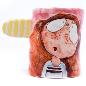 May include: A ceramic mug with a whimsical design. The mug features a cartoon-like face with red hair, freckles, and large eyes. The handle is shaped like a yellow and white striped ear.