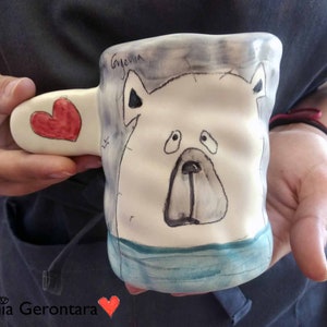Puede incluir: Una taza de cerámica blanca con un diseño pintado a mano de un oso polar con una franja azul y verde en la parte inferior. La taza tiene un asa en forma de corazón.