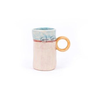 May include: A ceramic mug with a light blue, pink, and red glaze. The mug has a wooden handle and the words "Constar Goya" are written on the side.