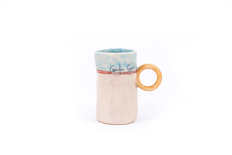 May include: A ceramic mug with a light blue, pink, and red glaze. The mug has a wooden handle and the words "Constar Goya" are written on the side.