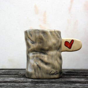 Puede incluir: Una taza de cer&aacute;mica con una superficie texturizada que se asemeja a la corteza de un &aacute;rbol. La taza tiene un asa con forma de rama y un coraz&oacute;n rojo pintado en el lateral. La taza est&aacute; firmada "Giaconda Grau".