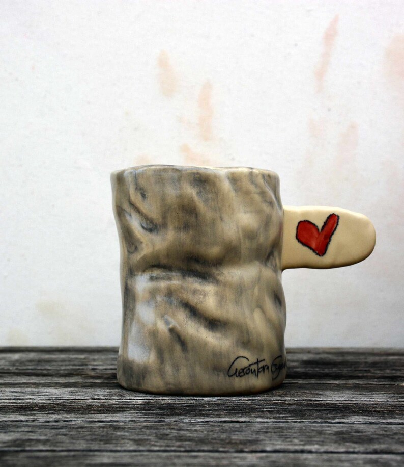 Puede incluir: Una taza de cer&aacute;mica con una superficie texturizada que se asemeja a la corteza de un &aacute;rbol. La taza tiene un asa con forma de rama y un coraz&oacute;n rojo pintado en el lateral. La taza est&aacute; firmada "Giaconda Grau".