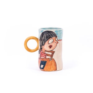 May include: A ceramic mug with a hand-painted design of a woman with black hair and a red and white striped shirt. The mug has a wooden handle and a light blue and beige background.