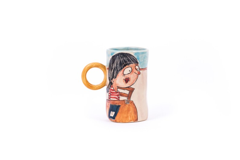 May include: A ceramic mug with a hand-painted design of a woman with black hair and a red and white striped shirt. The mug has a wooden handle and a light blue and beige background.