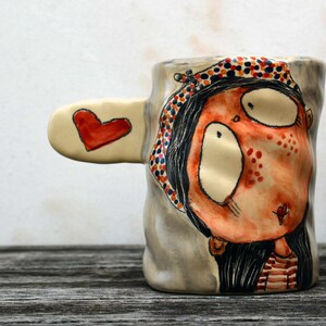 Puede incluir: Una taza de cer&aacute;mica con un dise&ntilde;o pintado a mano de una mujer con ojos grandes y un coraz&oacute;n rojo en la mejilla. La taza tiene un asa en forma de coraz&oacute;n y est&aacute; decorada con lunares rojos, naranjas y negros.