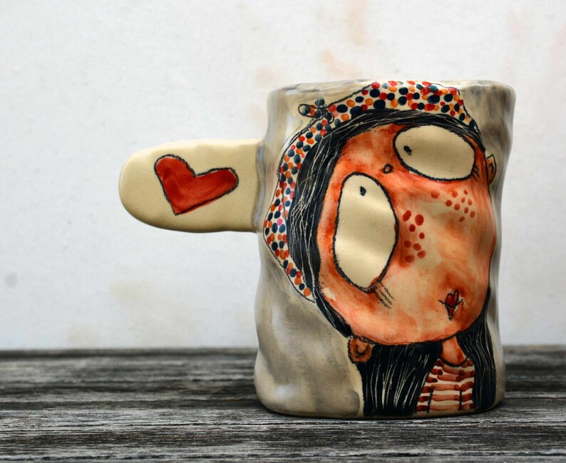 Puede incluir: Una taza de cer&aacute;mica con un dise&ntilde;o pintado a mano de una mujer con ojos grandes y un coraz&oacute;n rojo en la mejilla. La taza tiene un asa en forma de coraz&oacute;n y est&aacute; decorada con lunares rojos, naranjas y negros.