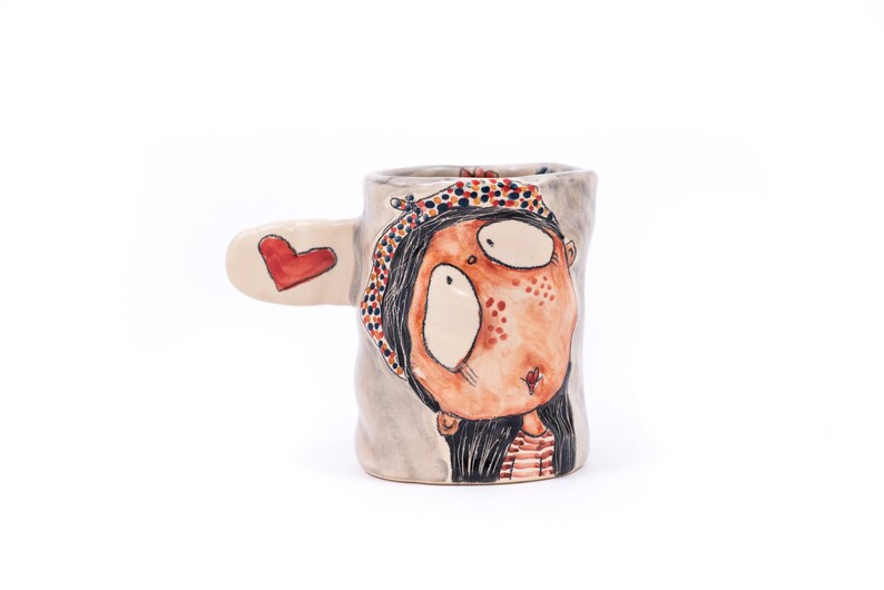Puede incluir: Una taza de cer&aacute;mica con un dise&ntilde;o pintado a mano de una ni&ntilde;a con un coraz&oacute;n rojo en el asa. La ni&ntilde;a tiene el pelo negro, un sombrero con estampado rojo y blanco y una camisa a rayas rojas y blancas.