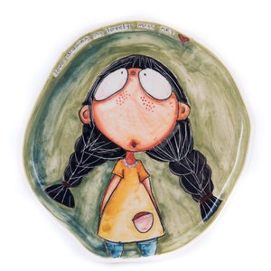 Puede incluir: Un plato de cerámica verde con una ilustración pintada a mano de una niña con cabello negro en trenzas. La niña lleva un vestido amarillo con un bolsillo rosa y pantalones vaqueros azules. El plato tiene el texto "love drawing my lovely miss art" escrito alrededor del borde.