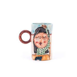 May include: A ceramic mug with a hand-painted design of a girl with closed eyes and a green and white polka dot shirt. The mug has a brown handle.