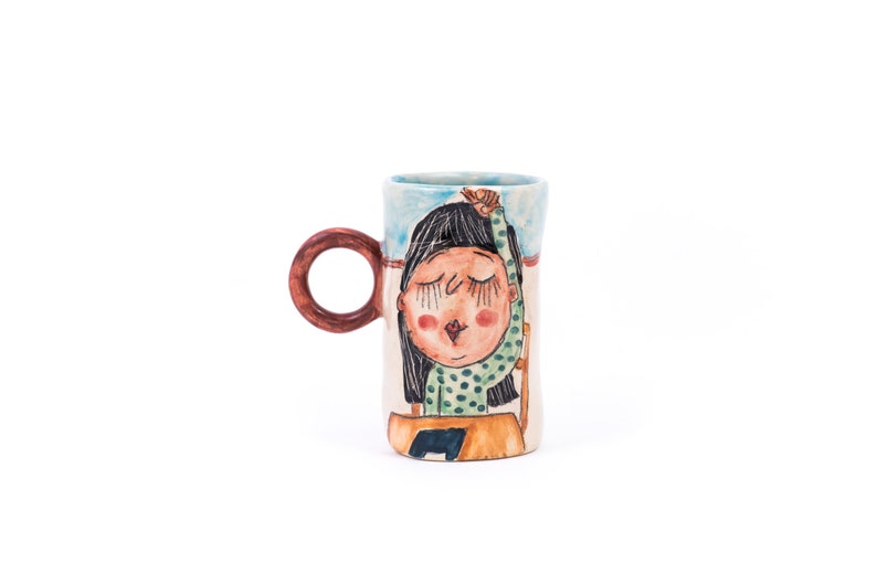 May include: A ceramic mug with a hand-painted design of a girl with closed eyes and a green and white polka dot shirt. The mug has a brown handle.