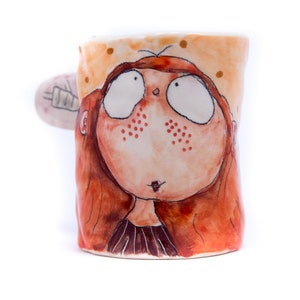 May include: A ceramic mug with a hand-painted design of a girl with red hair and freckles. The girl has large eyes and a surprised expression. The mug is painted in shades of orange, brown, and white.