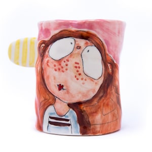 May include: A ceramic mug with a hand-painted design of a girl with brown hair, freckles, and big eyes. The mug has a pink background and a yellow and white striped handle.