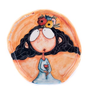 Puede incluir: Un plato de cerámica con una ilustración pintada a mano de una niña con ojos grandes y cabello negro. Lleva un vestido azul y sostiene una flor roja. El plato tiene un fondo naranja claro con el texto "Imagine you dream comes true" escrito en negro.