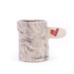 Puede incluir: Una taza de cer&aacute;mica con un esmalte marmoleado gris y blanco. La taza tiene un asa &uacute;nica con forma de rama de &aacute;rbol con un coraz&oacute;n rojo pintado en ella. La taza est&aacute; firmada "Geanita Bryan" en la parte inferior.