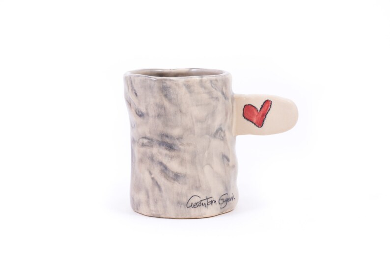 Puede incluir: Una taza de cer&aacute;mica con un esmalte marmoleado gris y blanco. La taza tiene un asa &uacute;nica con forma de rama de &aacute;rbol con un coraz&oacute;n rojo pintado en ella. La taza est&aacute; firmada "Geanita Bryan" en la parte inferior.