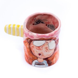 May include: A pink ceramic mug with a yellow and white striped handle. The mug is decorated with a cartoon illustration of a girl with red hair and freckles. The words "Gelateria Eugenia" are written on the side of the mug.