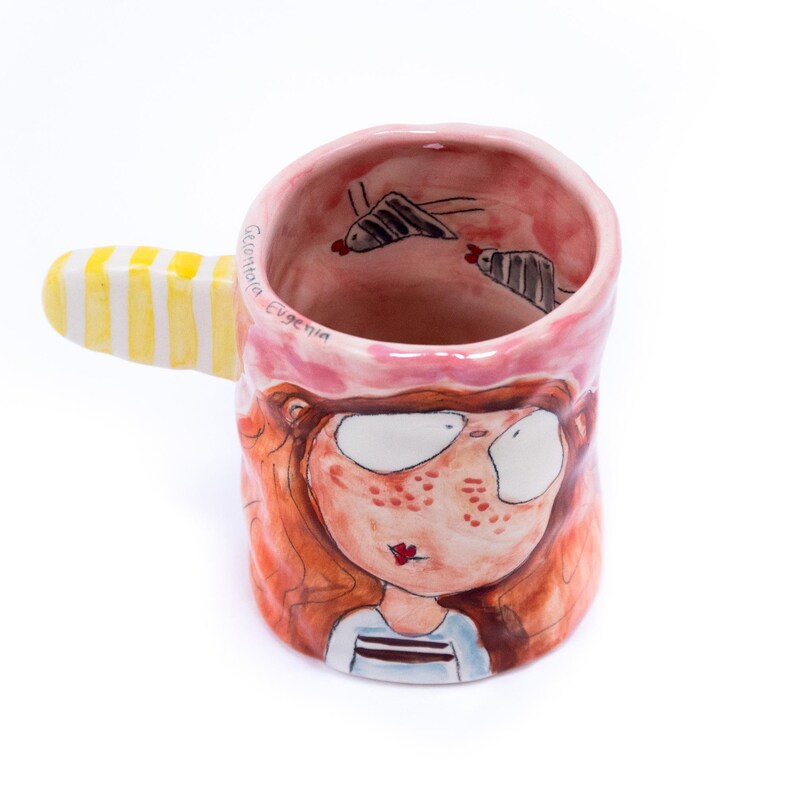 May include: A pink ceramic mug with a yellow and white striped handle. The mug is decorated with a cartoon illustration of a girl with red hair and freckles. The words "Gelateria Eugenia" are written on the side of the mug.