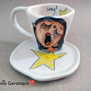 Op de afbeelding: Witte keramische koffiekop en schotel met een handgeschilderd ontwerp van een vrouw die zingt met haar mond open en haar ogen dicht. De kop heeft het woord "Sing!" erop geschreven. De schotel heeft een gele ster.