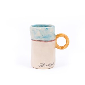 May include: A white ceramic mug with a blue and pink painted design and a yellow handle. The mug has the signature "Adrian Gray" on the bottom.
