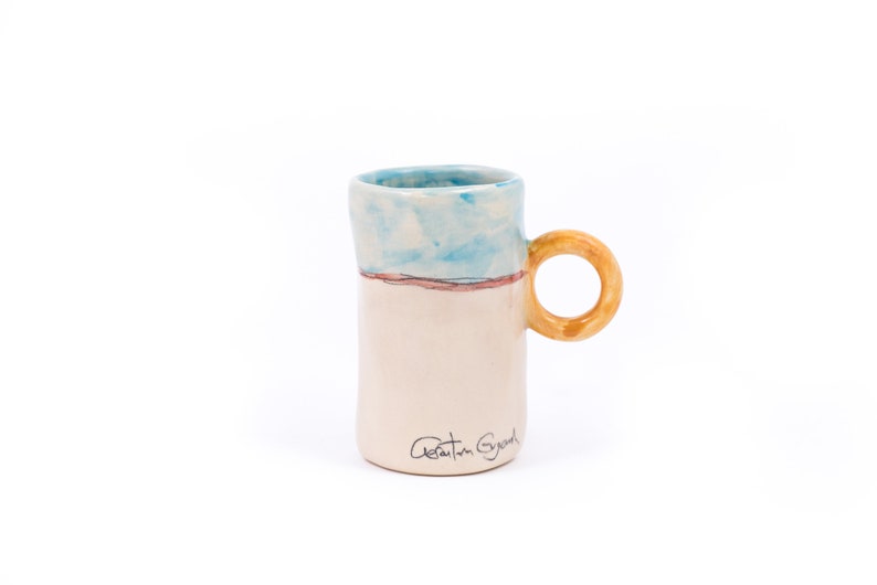 May include: A white ceramic mug with a blue and pink painted design and a yellow handle. The mug has the signature "Adrian Gray" on the bottom.