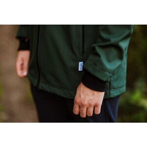 Puede incluir: Una chaqueta softshell verde oscuro con cierre de cremallera y una peque&ntilde;a etiqueta en el lado izquierdo. La chaqueta tiene un forro interior negro y un pu&ntilde;o negro en la manga izquierda.