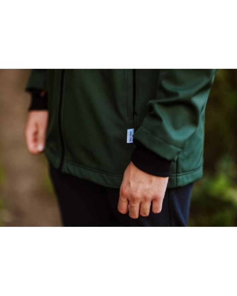 Puede incluir: Una chaqueta softshell verde oscuro con cierre de cremallera y una peque&ntilde;a etiqueta en el lado izquierdo. La chaqueta tiene un forro interior negro y un pu&ntilde;o negro en la manga izquierda.
