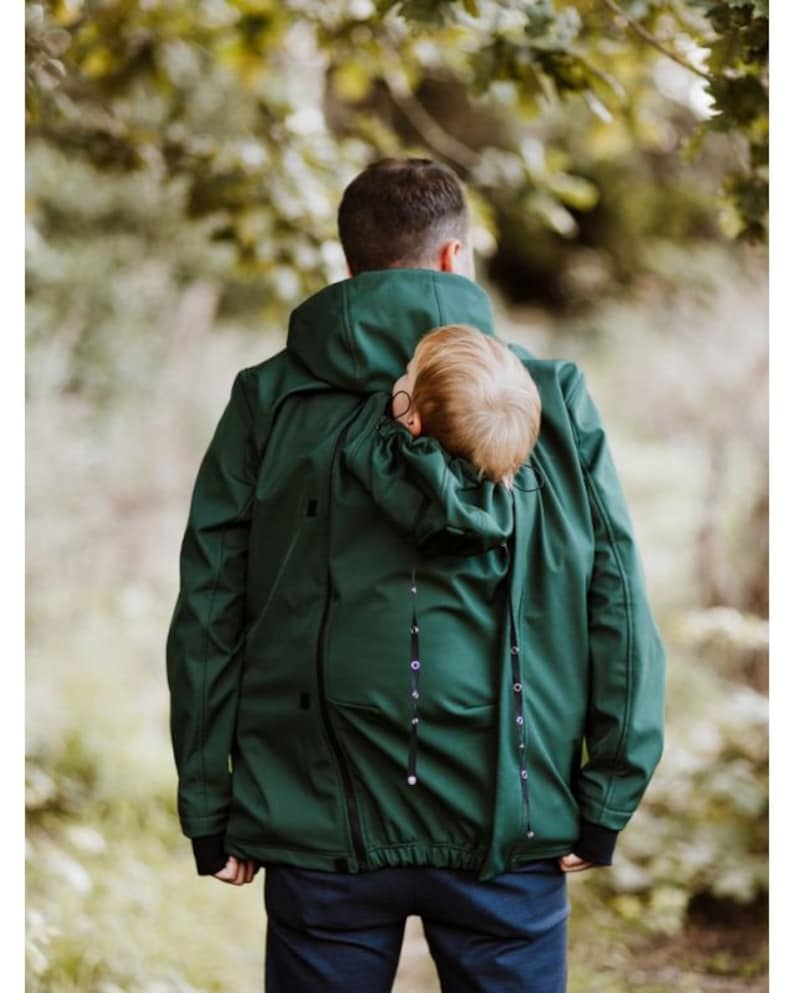 Puede incluir: Una chaqueta verde oscuro con un portabeb&eacute;s integrado. La chaqueta tiene una capucha y una cremallera. El portabeb&eacute;s es visible a trav&eacute;s de la parte posterior de la chaqueta.