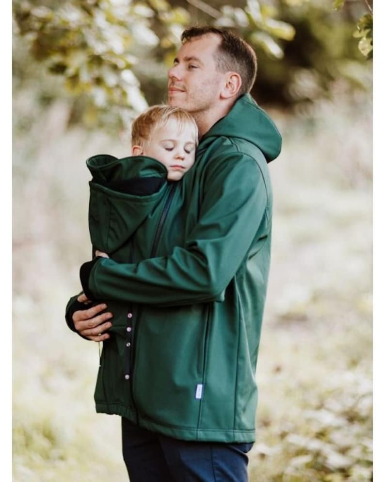 Puede incluir: Una chaqueta verde oscuro con capucha, cierre de cremallera y un portabeb&eacute;s integrado. La chaqueta tiene una cremallera negra y una peque&ntilde;a etiqueta blanca en la parte inferior derecha.