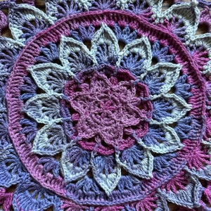 May include: A crocheted mandala with a purple, lavender, and white color scheme. The mandala is made with a variety of stitches and has a delicate, intricate design.