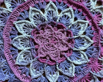 May include: A crocheted mandala with a purple, lavender, and white color scheme. The mandala is made with a variety of stitches and has a delicate, intricate design.