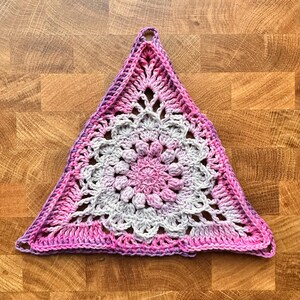 Op de afbeelding: Een handgemaakte, driehoekige gehaakte doily in tinten roze, paars en grijs. De doily heeft een centraal bloemmotief en een open patroon. Er zit een lus in de bovenhoek om op te hangen. De doily is geplaatst op een houten oppervlak.