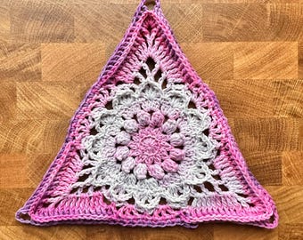 May include: A handmade, triangular crochet doily in shades of pink, purple, and gray. The doily features a central floral design and an openwork pattern. A loop is at the top corner for hanging. The doily is placed on a wooden surface.