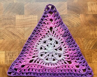 May include: A triangular crochet motif featuring a gradient color pattern that transitions from a light lavender center to vibrant pink and deep purple hues at the edges. The design incorporates an intricate openwork stitch pattern.