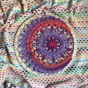 Op de afbeelding: Een paarse en oranje gehaakte mandala met een houten hoepel, op een wit en kleurrijk gehaakt deken.
