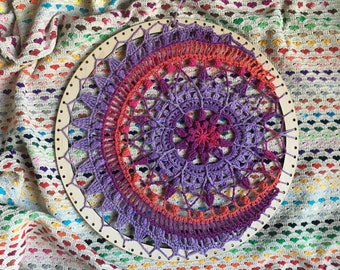 May include: A purple and orange crochet mandala with a wooden hoop, on a white and colorful crocheted blanket.