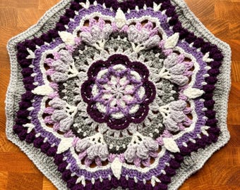May include: A hexagonal crochet doily featuring a mandala pattern in shades of purple, lavender, white, and gray. The design includes intricate floral-like clusters, textured bobble stitches, and a scalloped border, all worked in yarn.