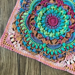 Könnte beinhalten: Ein farbenfroher gehäkelter Quadrat mit einem Mandala-Design in den Farben Rosa, Grün, Blau und Braun. Das Quadrat ist mit verschiedenen Maschen und Texturen gearbeitet, wodurch ein einzigartiges und filigranes Muster entsteht.