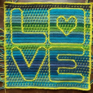 May include: A square, handmade crochet piece with the word "LOVE" and a heart symbol. The letters and heart are outlined in yellow, with a variety of blue and green shades filling the spaces. The edges are trimmed with yellow yarn.
