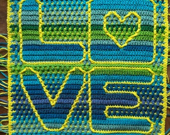 May include: A square, handmade crochet piece with the word "LOVE" and a heart symbol. The letters and heart are outlined in yellow, with a variety of blue and green shades filling the spaces. The edges are trimmed with yellow yarn.