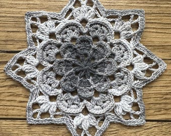 May include: A gray and white crocheted star-shaped doily with a detailed design.