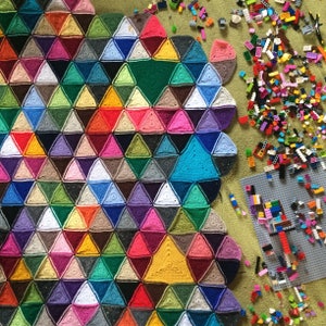 May include: A colorful crocheted blanket with a scalloped edge, made of various shades of pink, blue, green, yellow, and brown. The blanket is made of triangular stitches and is laid out on a green surface.