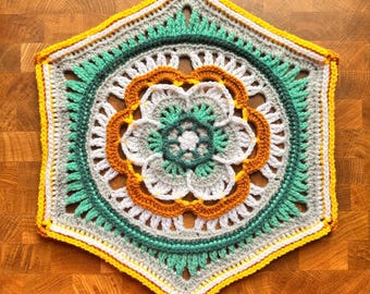 May include: A hexagonal crochet motif featuring a central white flower surrounded by layers of light gray, teal, and orange yarn. The design includes intricate stitch patterns with a bright yellow border, creating a geometric floral textile piece.