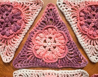 May include: Three triangular crochet motifs featuring a central floral pattern in shades of pink, purple, and white. Each piece is crafted with yarn in a gradient color scheme, showcasing intricate openwork stitches and a textured flower center.
