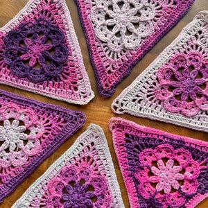 Op de afbeelding: Vijf driehoekige haakmotieven met een centraal bloempatroon in tinten paars, magenta en wit. Elk stuk toont een ingewikkeld opengewerkt ontwerp met een geschulpte rand, met een kleurverloop van licht naar donker in het garen.