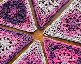 May include: Five triangular crochet motifs featuring a central floral pattern in shades of purple, magenta, and white. Each piece displays an intricate openwork design with a scalloped edge, showcasing a gradient of yarn colors from light to dark.