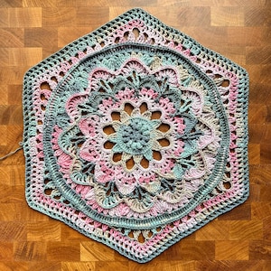 Puede incluir: Un tapete de ganchillo hecho a mano con forma hexagonal. El tapete presenta un diseño floral con círculos concéntricos de calados y puntos de ganchillo sólidos. La paleta de colores incluye tonos de rosa, verde y beige, creando una estética suave y pastel.