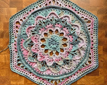 May include: A handmade, crocheted doily in a hexagonal shape. The doily features a floral design with concentric circles of openwork and solid crochet stitches. The color palette includes shades of pink, green, and beige, creating a soft, pastel aesthetic.