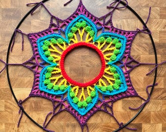 May include: A vibrant, handmade crochet dreamcatcher featuring a central flower design. The flower has a red center, surrounded by yellow, green, and blue petals, all set against a purple background. The dreamcatcher is suspended from a black metal ring.